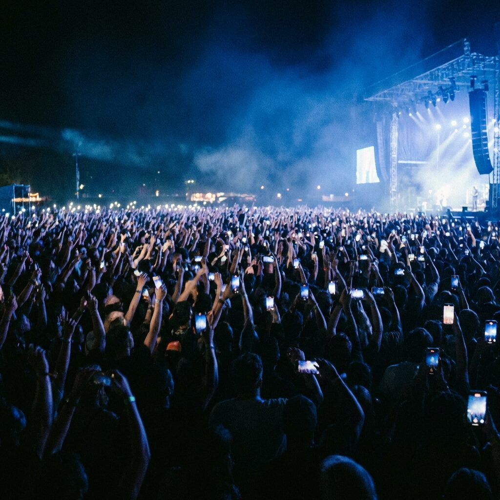 A massive outdoor festival crowd at night, thousands of people with hands raised and phone lights glowing, illuminated by stage light in deep blue and white. The perspective is from behind the crowd looking toward a distant brightly lit stage. The visual conveys the specific electricity of collective singing — an enormous number of people united in a single shared moment. Cinematic, atmospheric, slightly blurred motion in the crowd to suggest energy and movement. No identifiable faces.